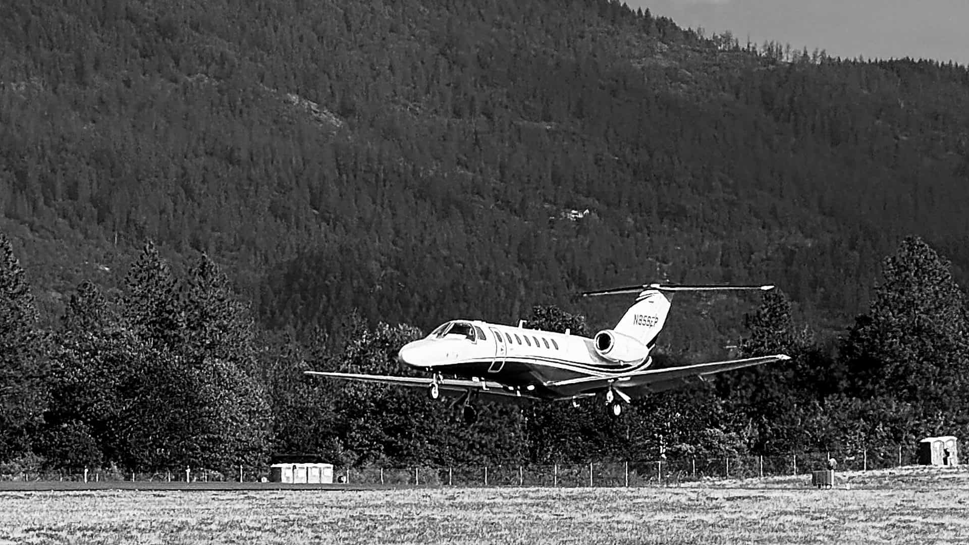 A twin engine business jet on short final, a forested hillside rising behind it.