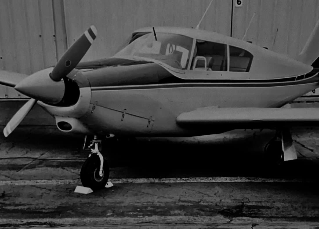 A Piper Comanche parked in front of a hangar, photographed in black and white.