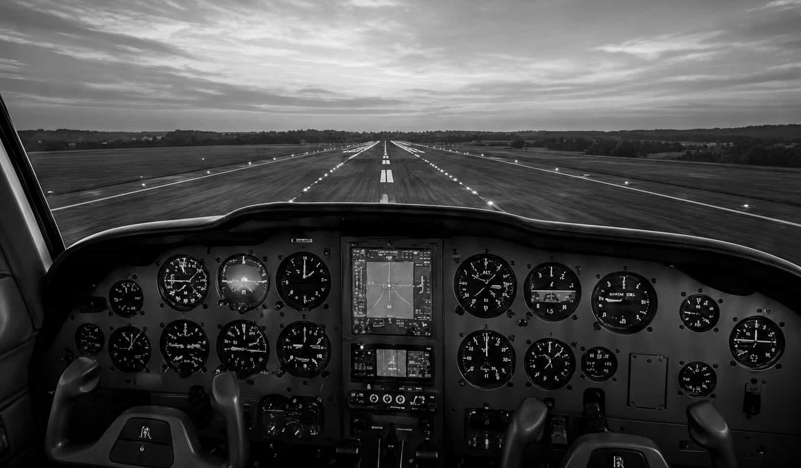 The view forward over a light aircraft's instrument panel and down a lit runway at sunset.