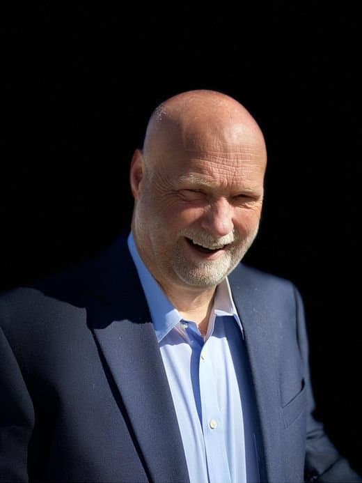 Jim Carpenter in a navy jacket, laughing, lit from one side against a dark background.